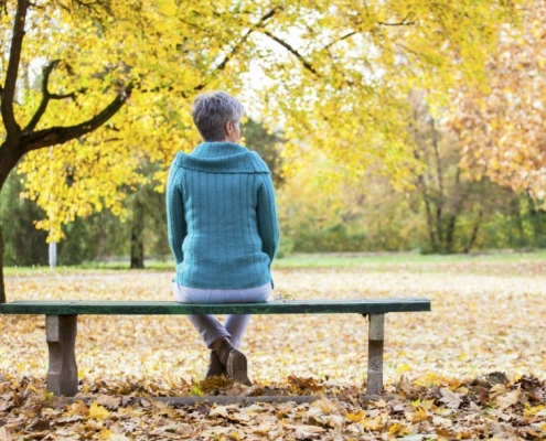 A lady sitting on a park bench with autumn leaves behind her.