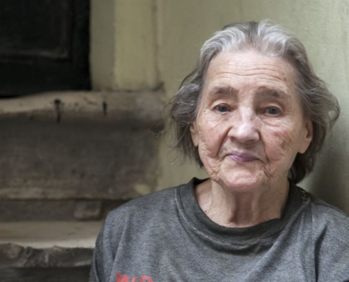 An older lady in a grey top, she is sitting on a stairwell to a building