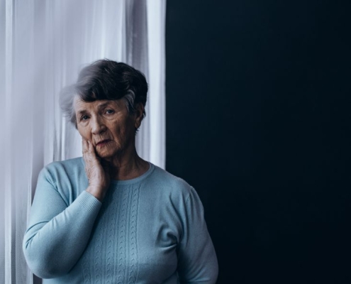 And older woman standing in front of net blinds with her hand to her face and a worried look.