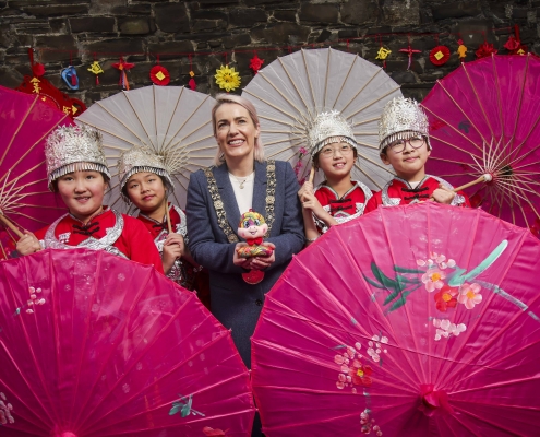 4 girls in traditional chinese clothes while the lord mayor of Dublin poses with them, all have smiles on their faces and pink umbrellas are in the foreground and background.