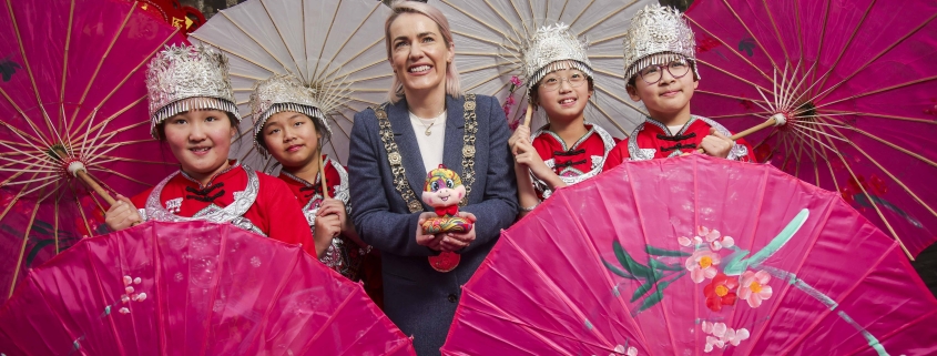 4 girls in traditional chinese clothes while the lord mayor of Dublin poses with them, all have smiles on their faces and pink umbrellas are in the foreground and background.